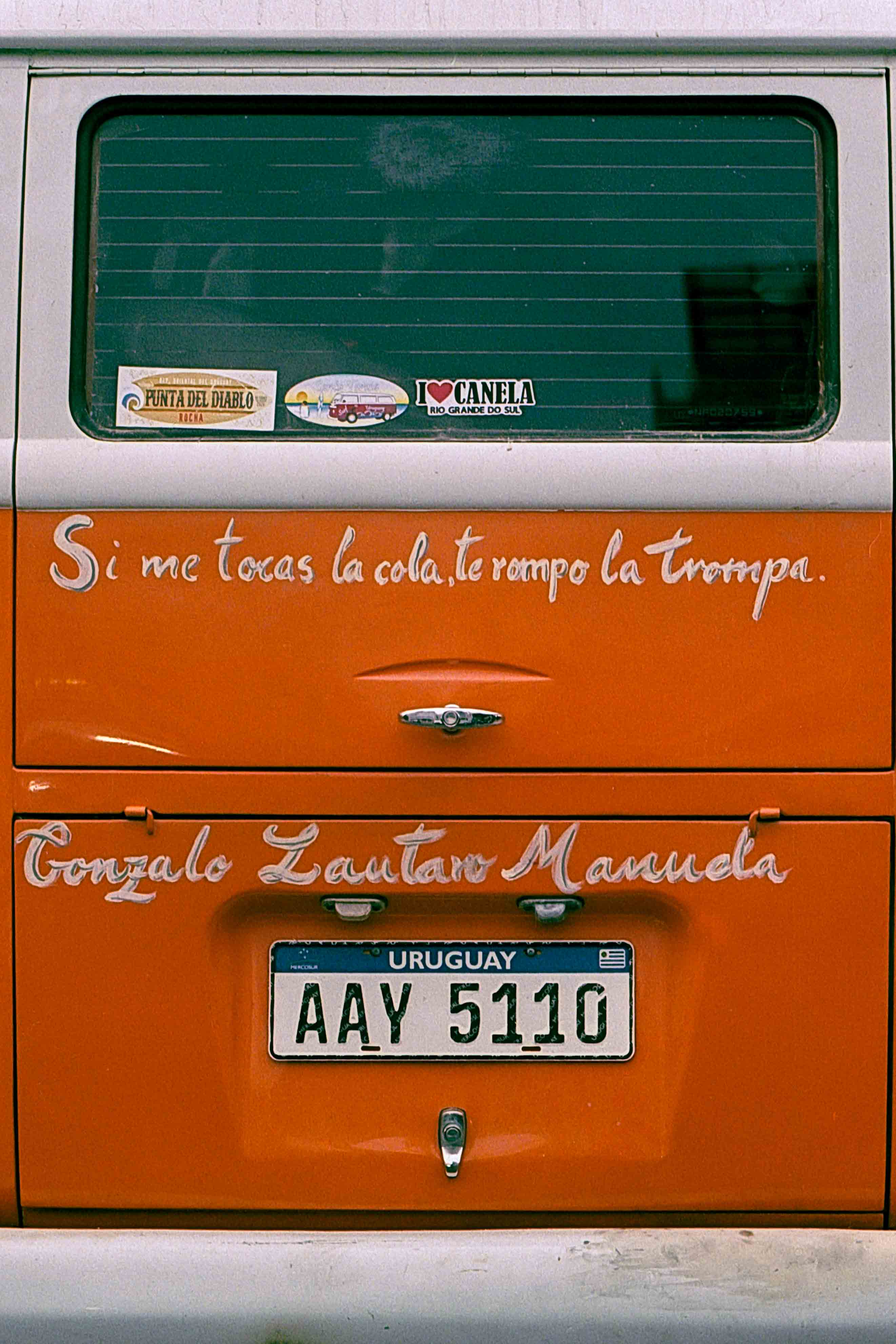 Puertas traseras naranjas de una furgoneta con una frase y nombres pintados en blanco.