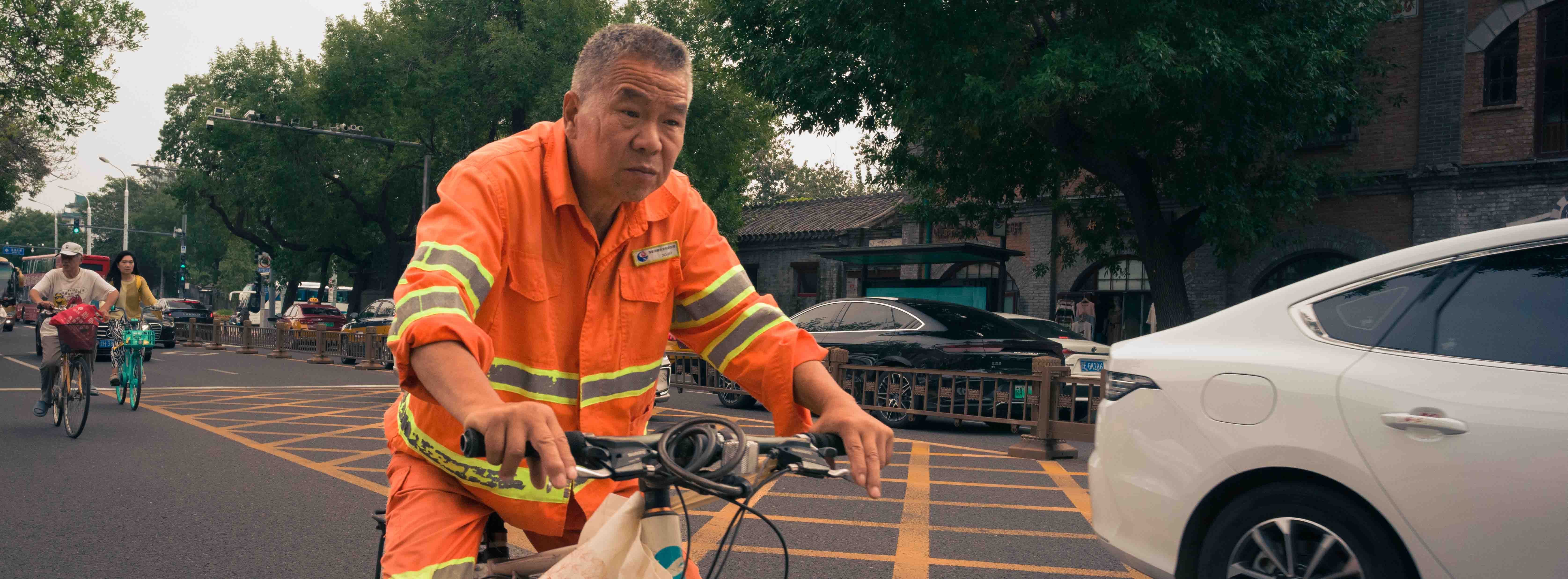 Persona con uniforme naranja y franjas reflectantes conduce una bicicleta junto al tráfico.