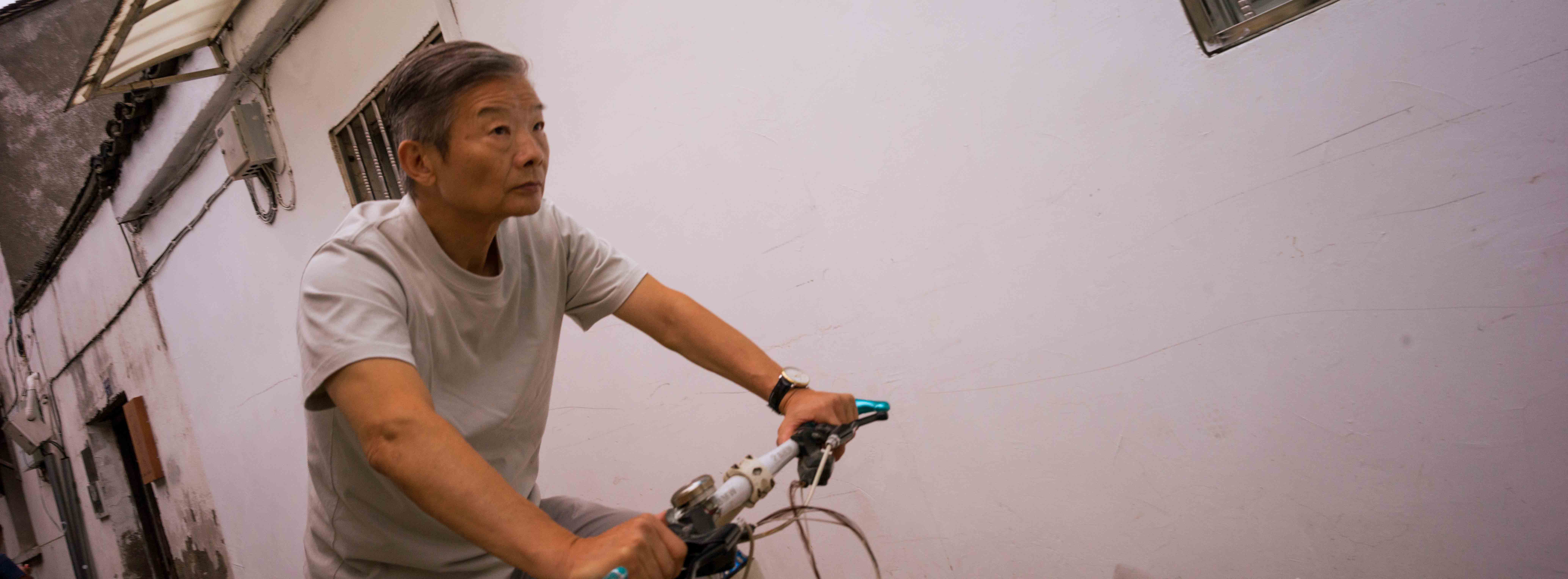 Persona con camiseta gris conduce una bicicleta junto a una pared blanca.