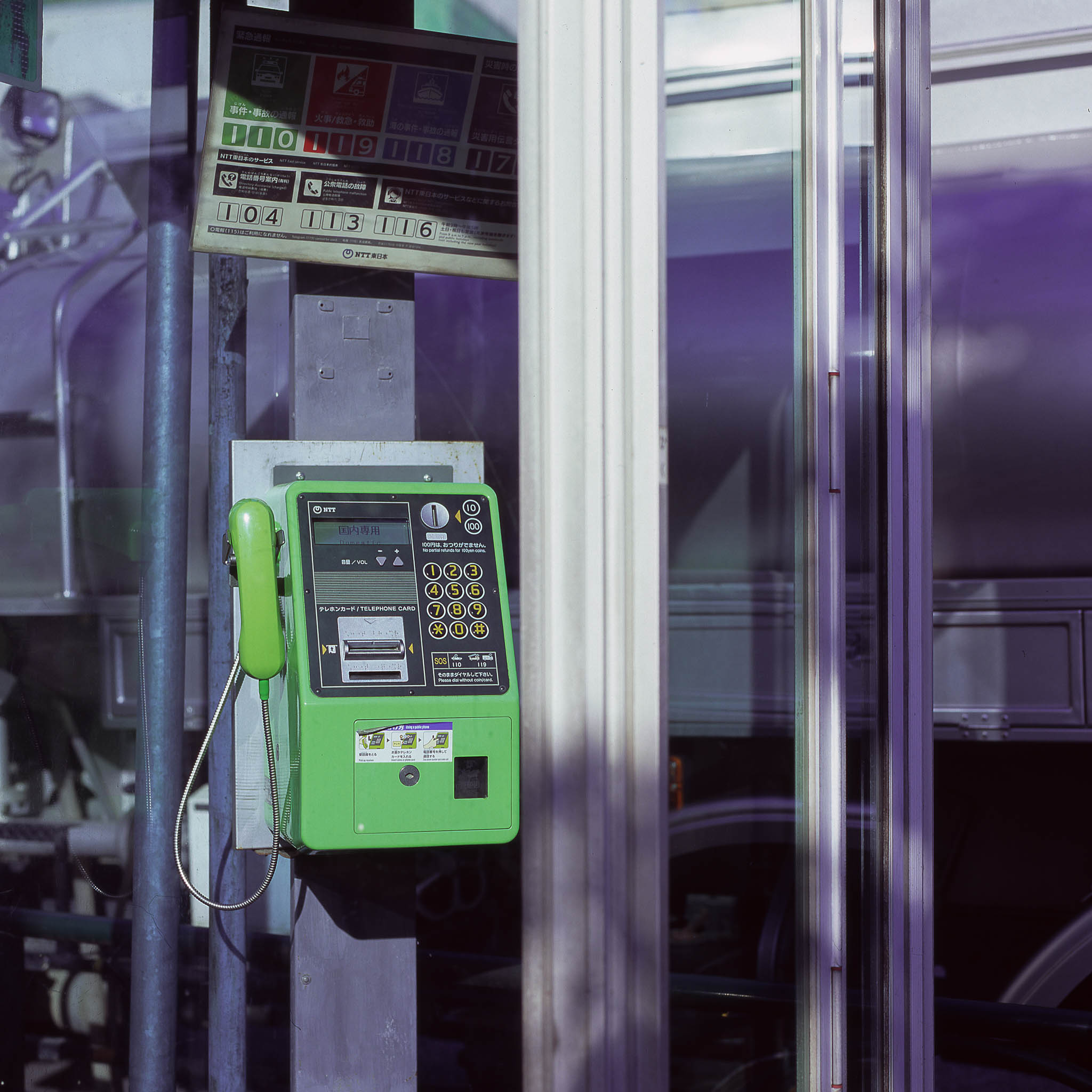 Teléfono público verde dentro de una cabina de vidrio, bajo un cartel con números de servicio.