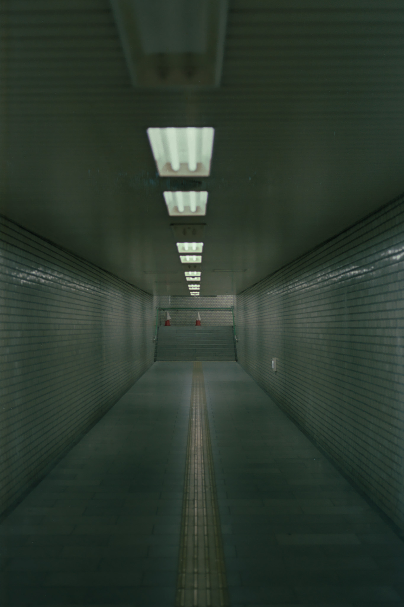 Pasillo vacío con paredes de azulejos, luces en el techo y una franja táctil que conduce a unas escaleras.
