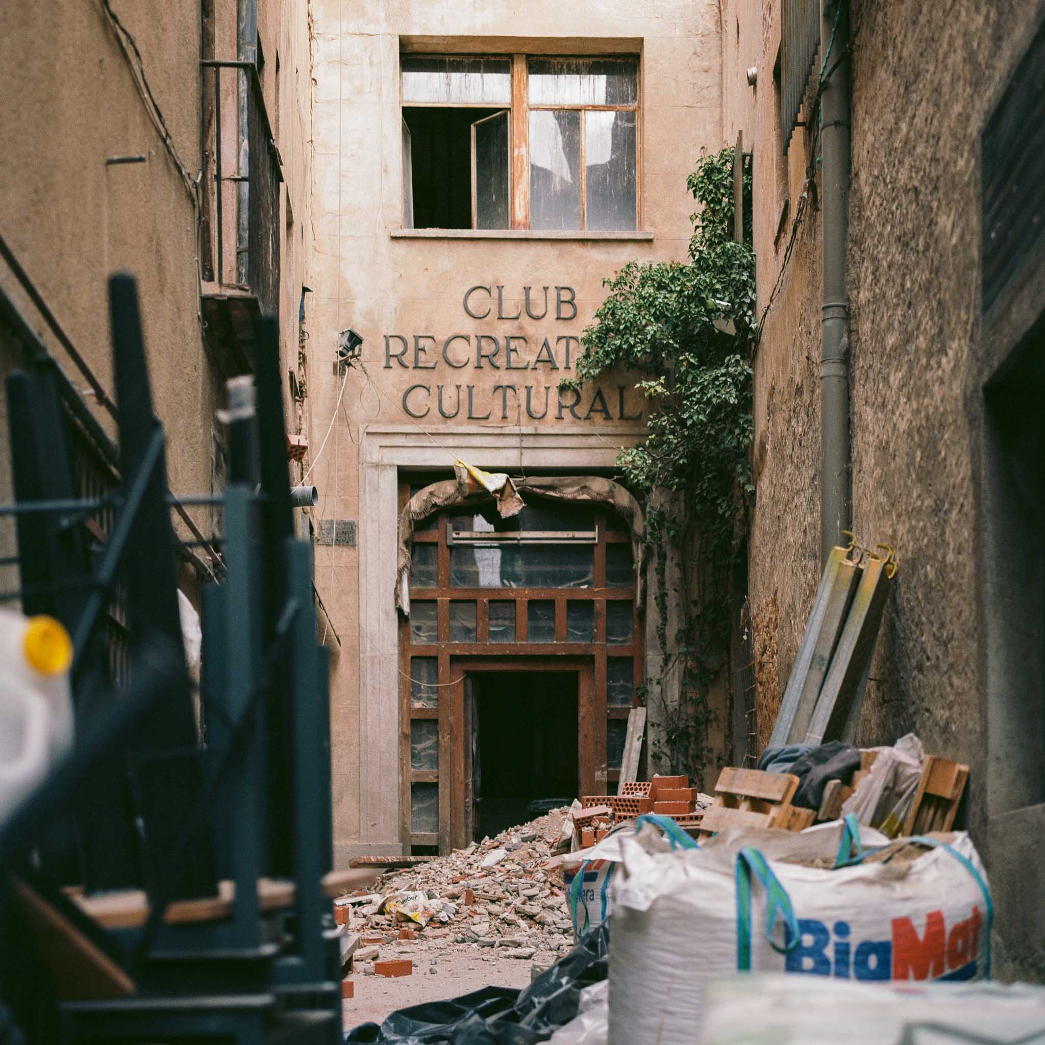Entrada deteriorada de un club cultural, con escombros, bolsas de obra y una enredadera en la fachada.