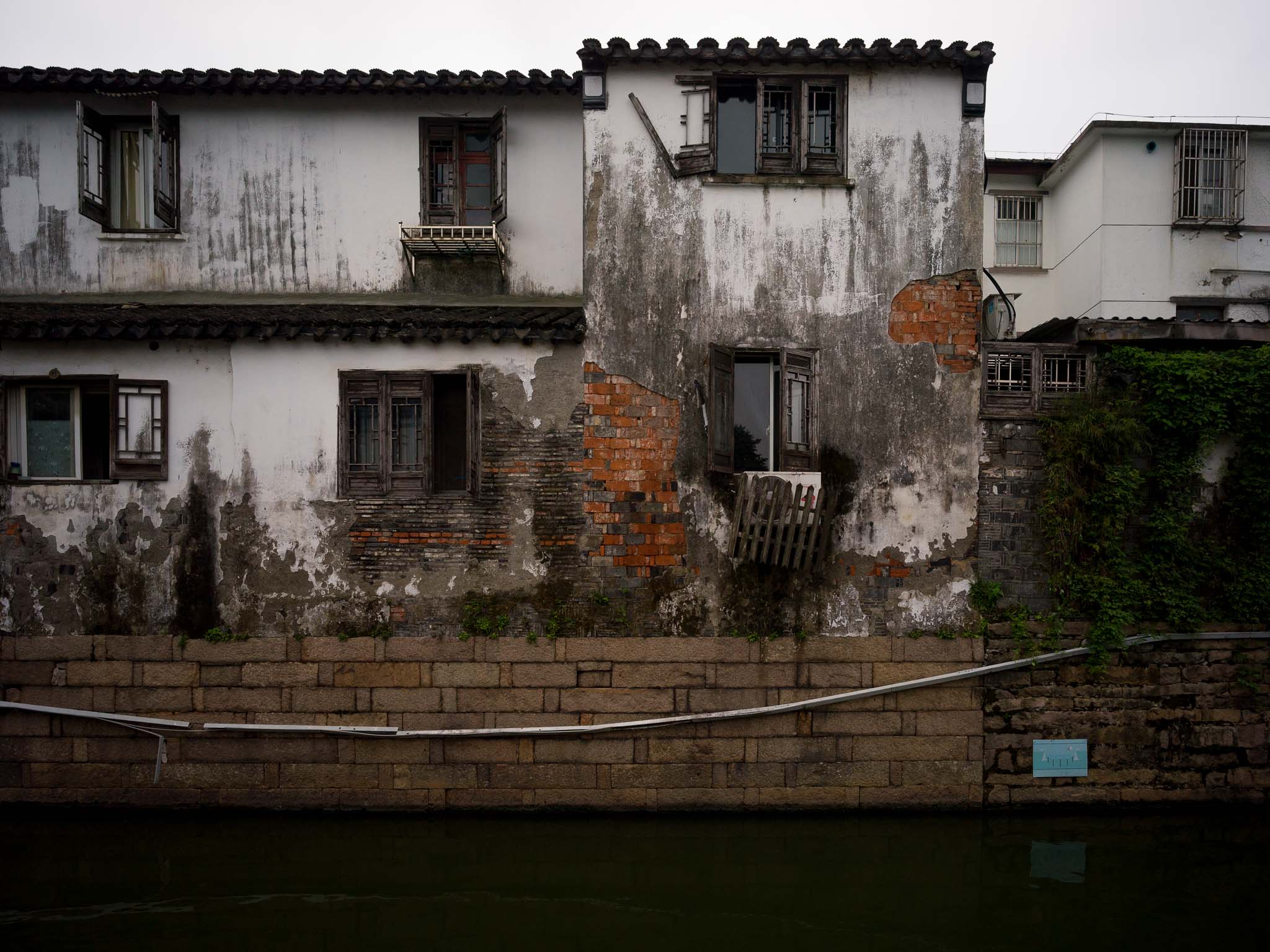 Fachada junto al agua con revoque descascarado, ladrillos a la vista y ventanas de madera abiertas.