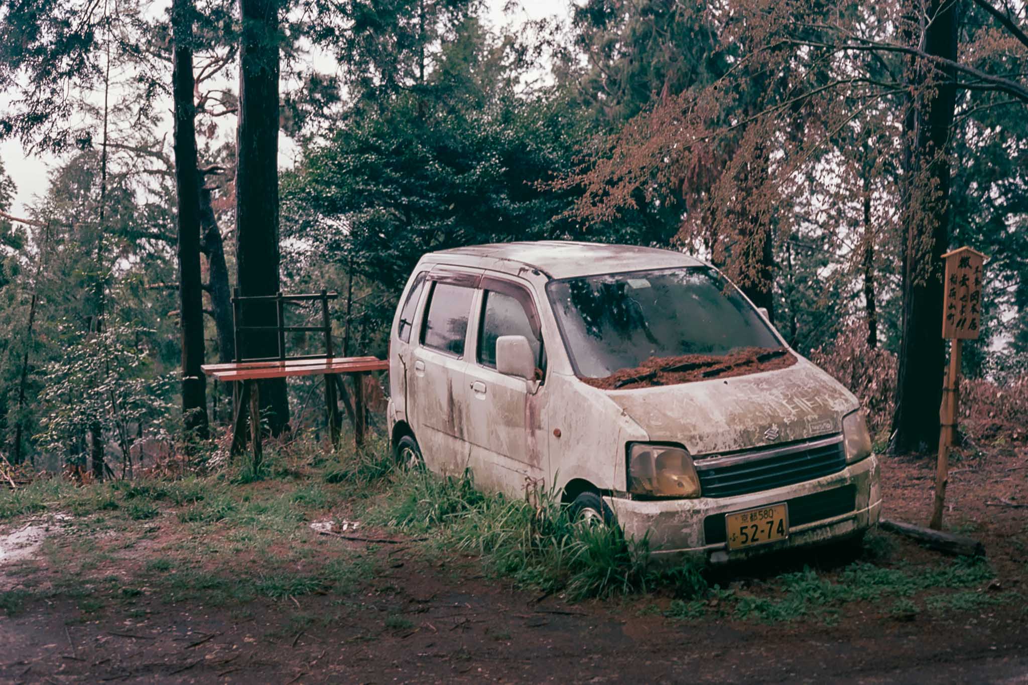 Automóvil blanco cubierto de suciedad y hojas, estacionado entre árboles con hierba alrededor de las ruedas.