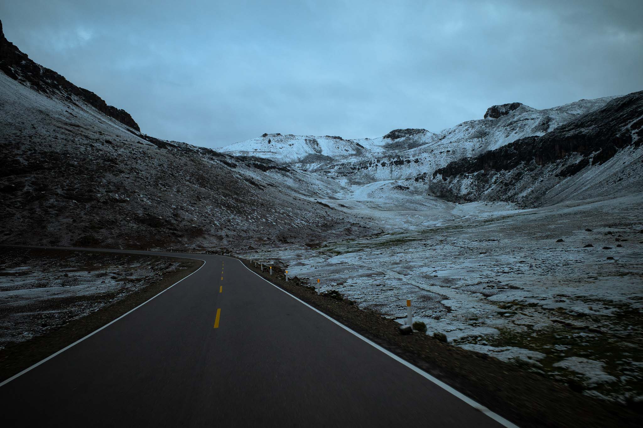 Ruta vacía que se curva entre montañas rocosas parcialmente cubiertas de nieve, bajo un cielo nublado.