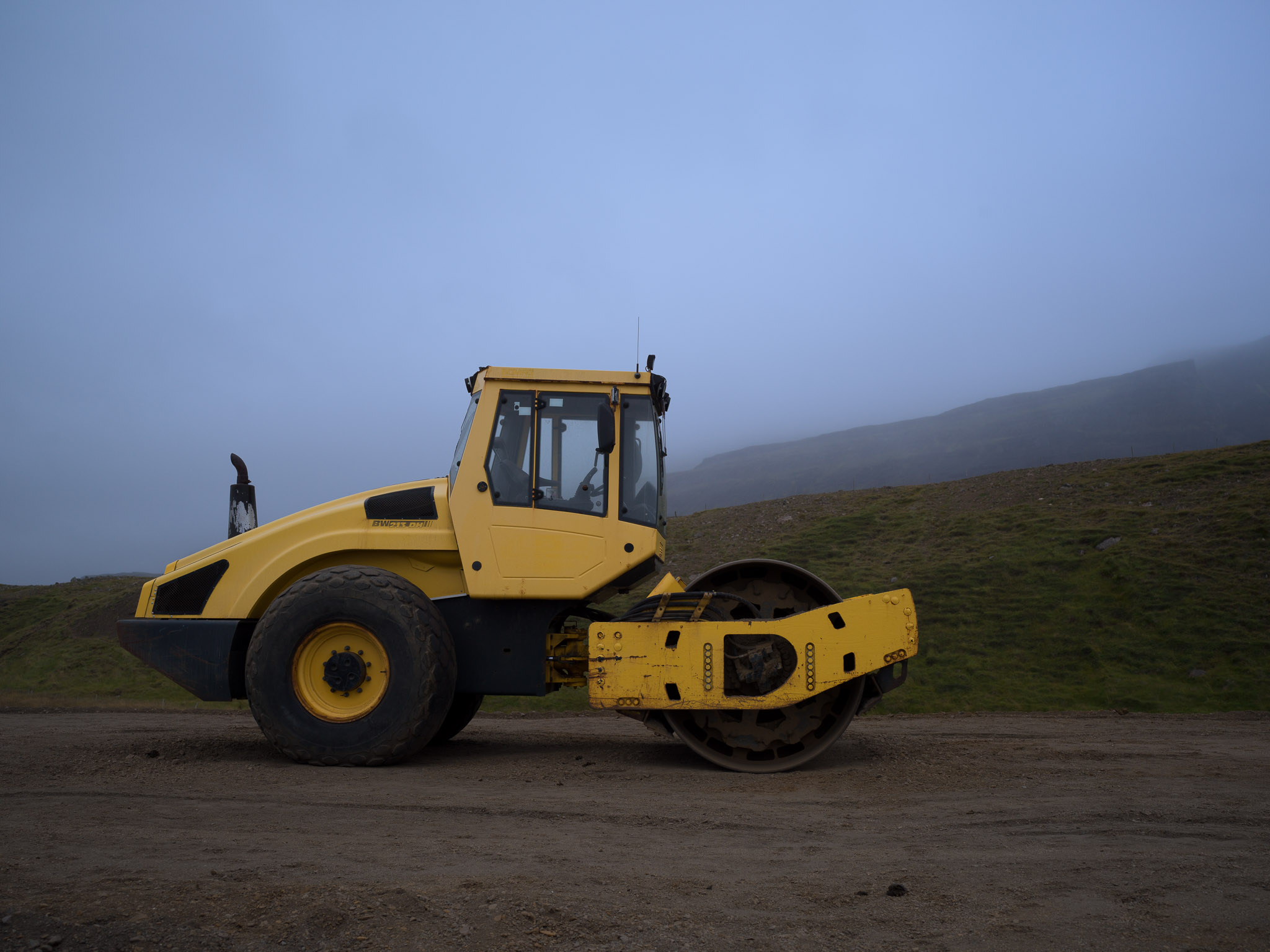 Rodillo compactador amarillo estacionado sobre tierra junto a una ladera cubierta de pasto y niebla.