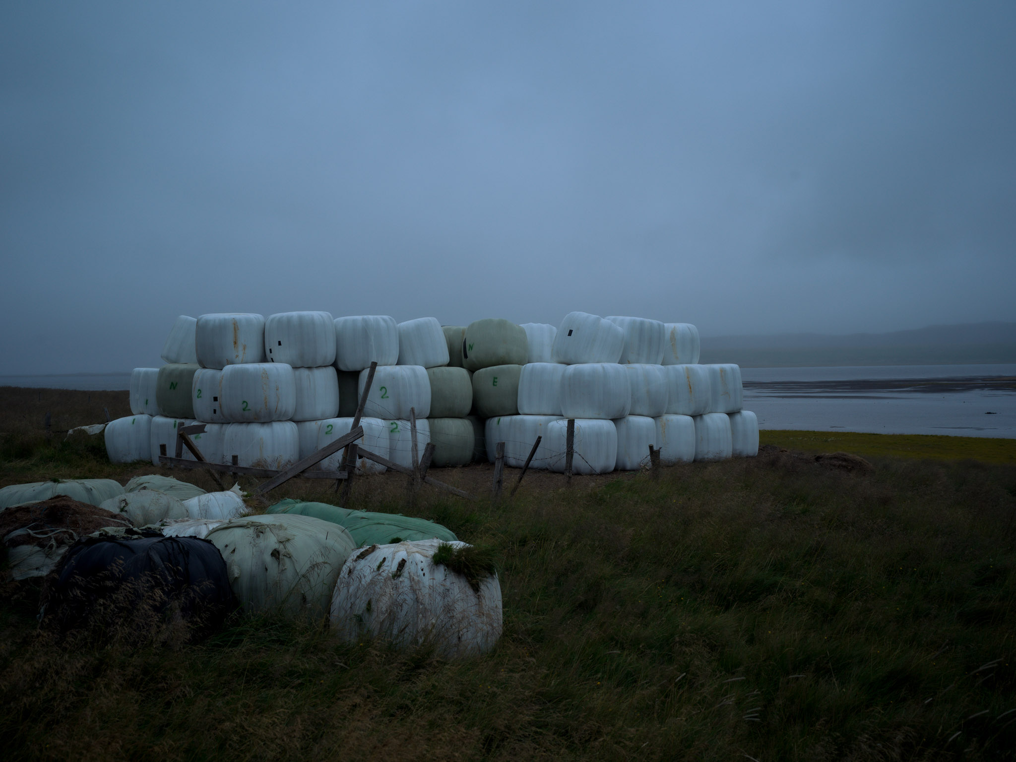 Rollos envueltos en plástico blanco apilados junto al agua, detrás de un cerco de madera deteriorado, entre la niebla.