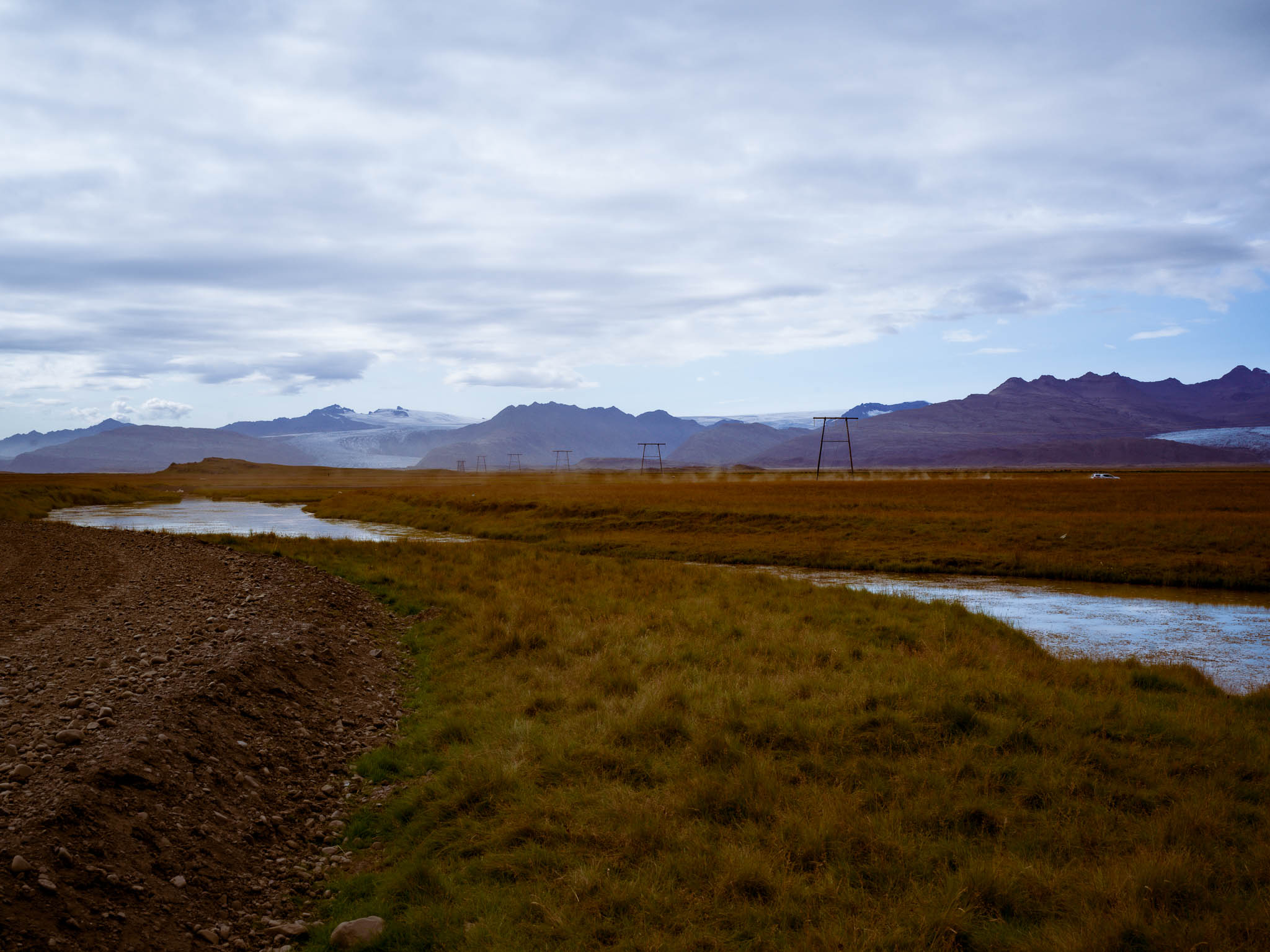 Arroyo sinuoso entre pastizales dorados, con torres eléctricas y montañas con glaciares al fondo.