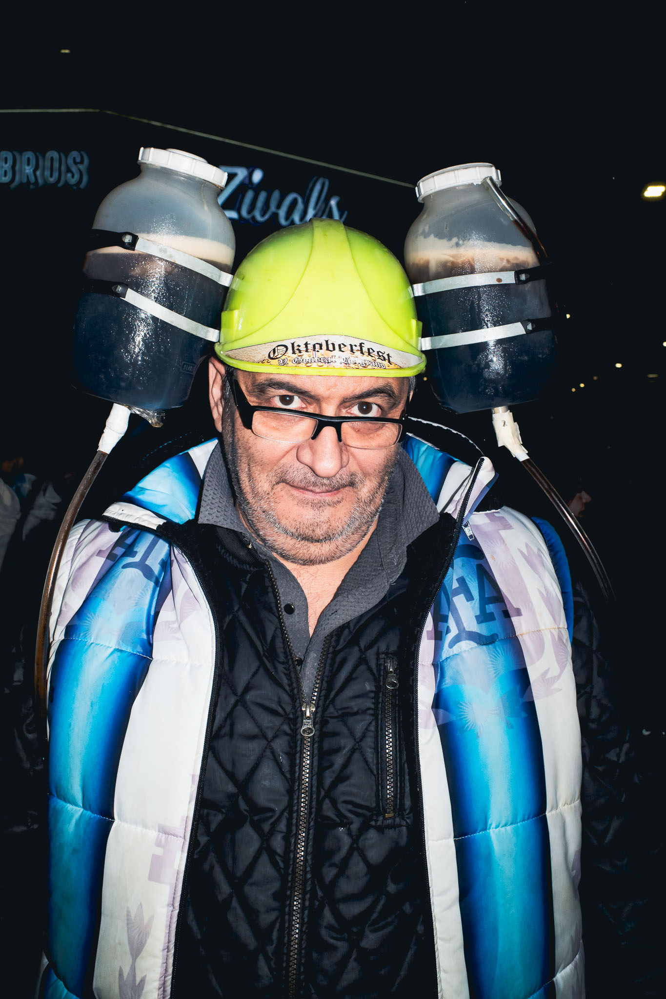 Persona con casco verde y dos recipientes de bebida sujetos a los lados, vestida con una capa celeste y blanca sobre una campera negra.