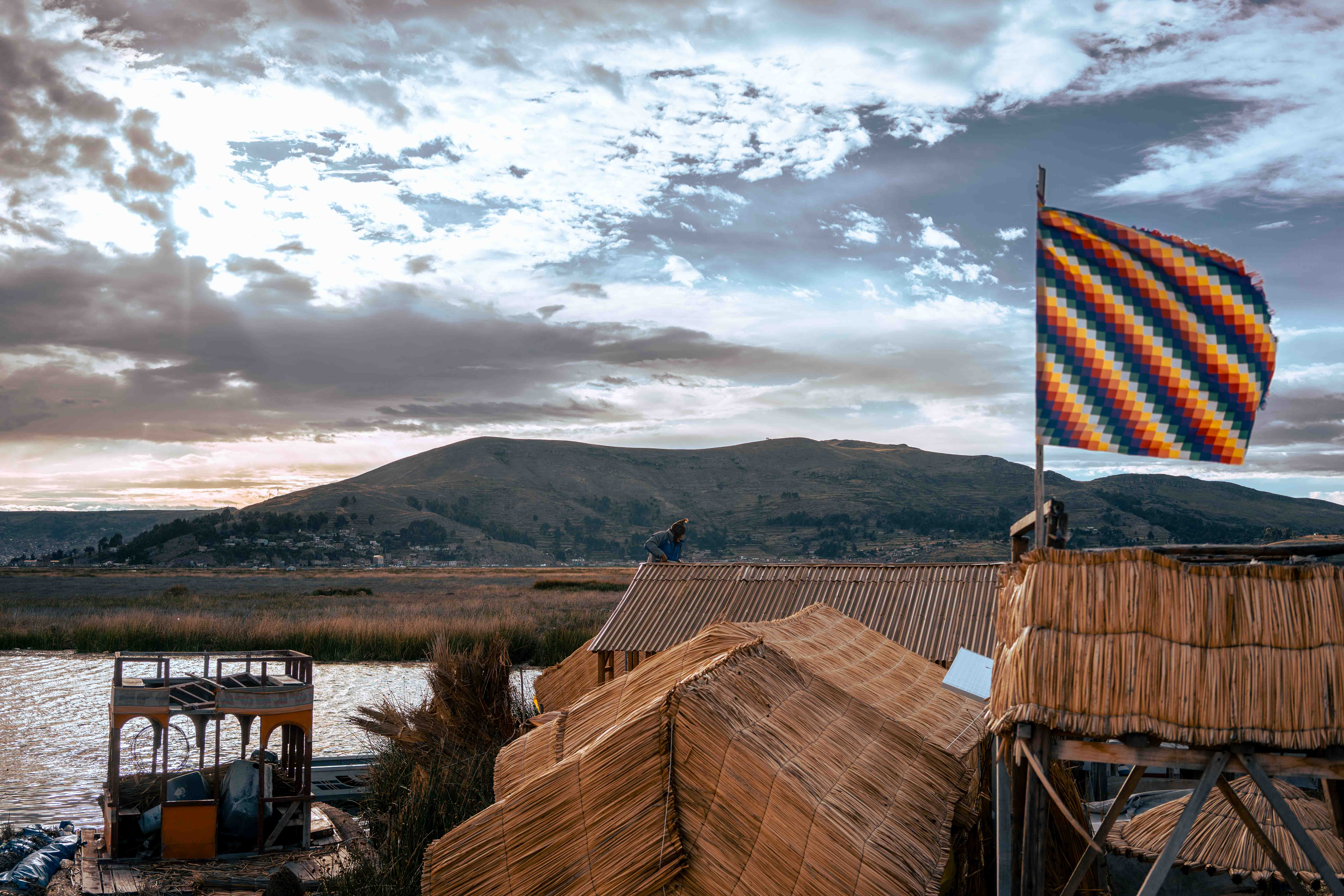 Bandera multicolor sobre techos de juncos, con agua y montañas bajo el sol entre nubes.