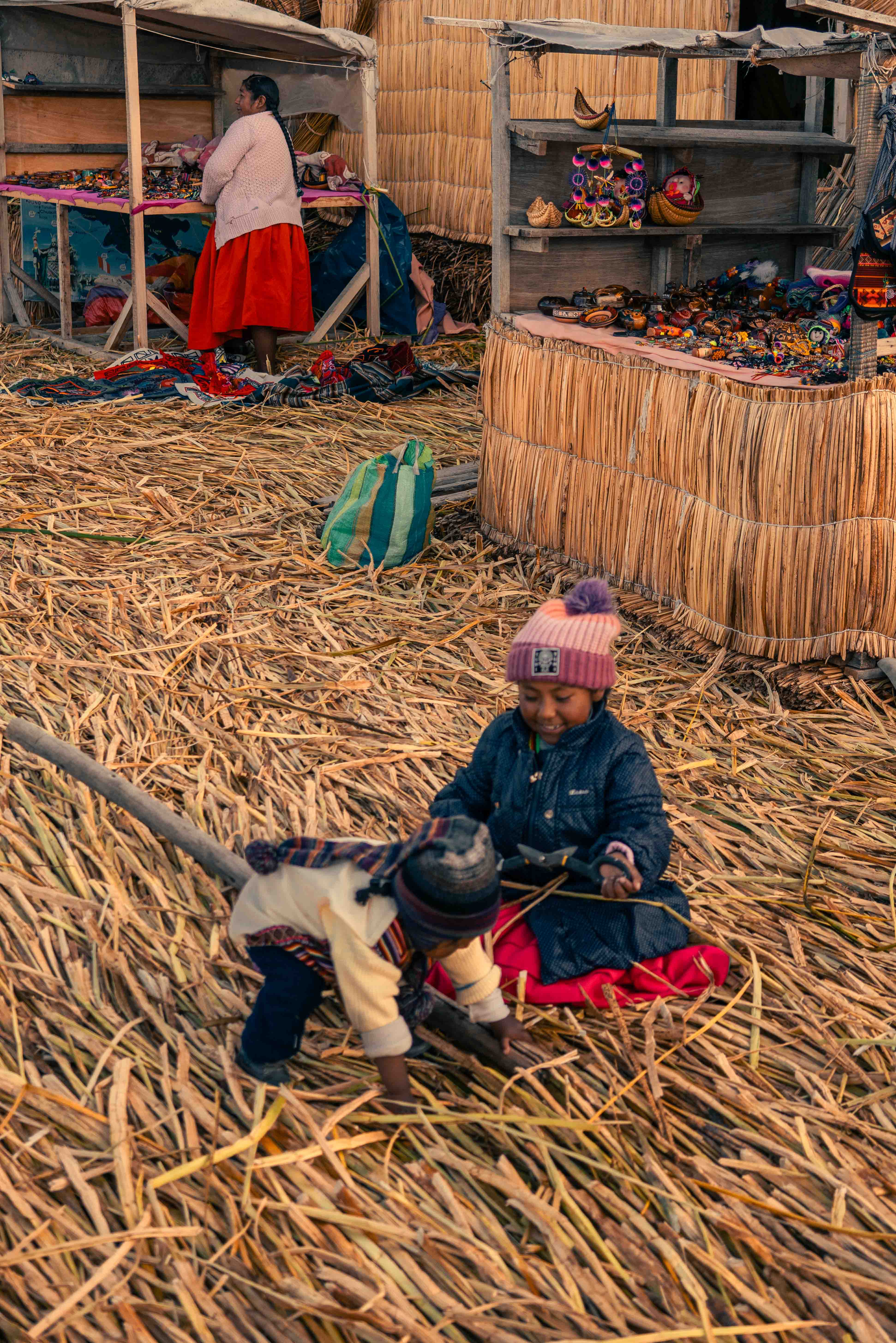 Figura pequeña con gorro sentada junto a objetos sobre un suelo de juncos, delante de un puesto.