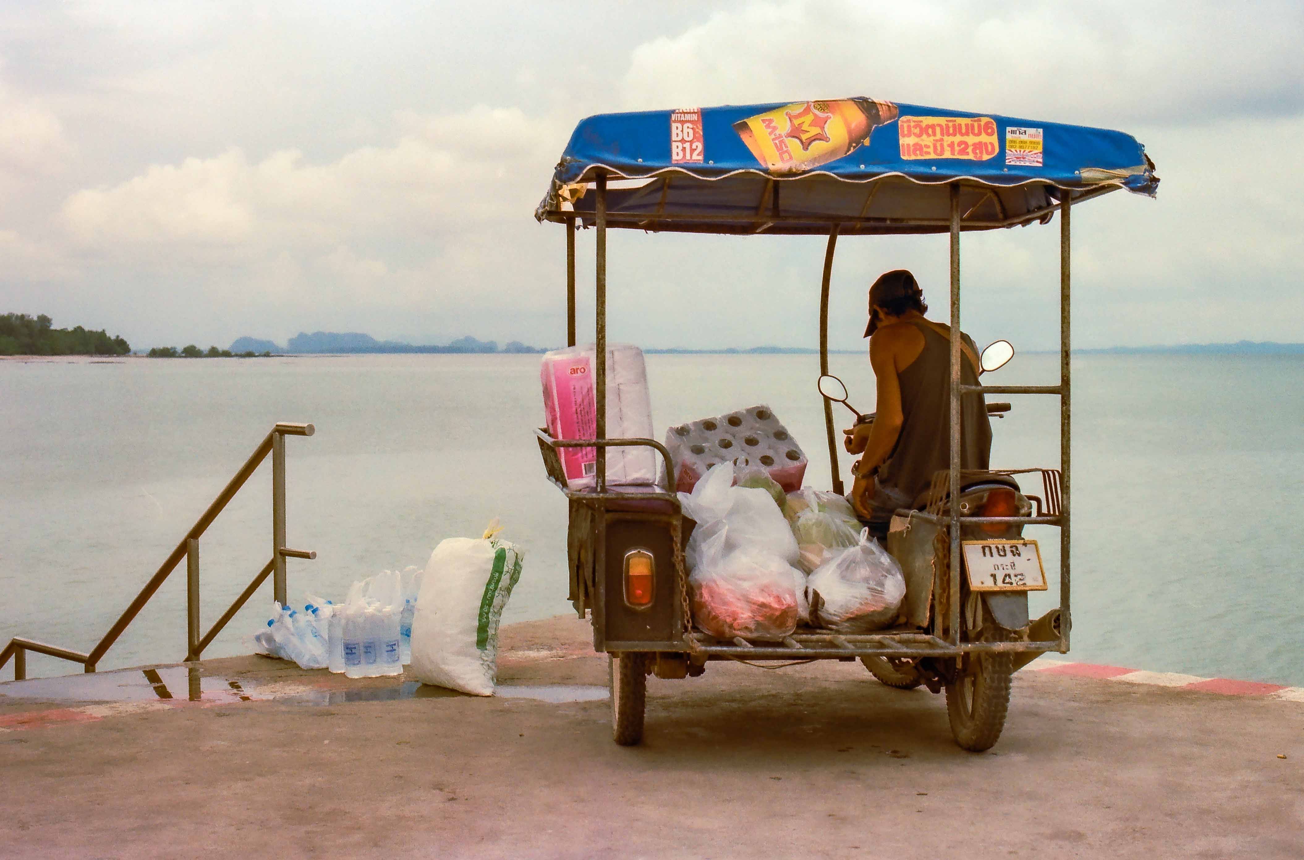 Persona junto a un carrito de venta con toldo azul, bolsas y botellas a la orilla del agua.