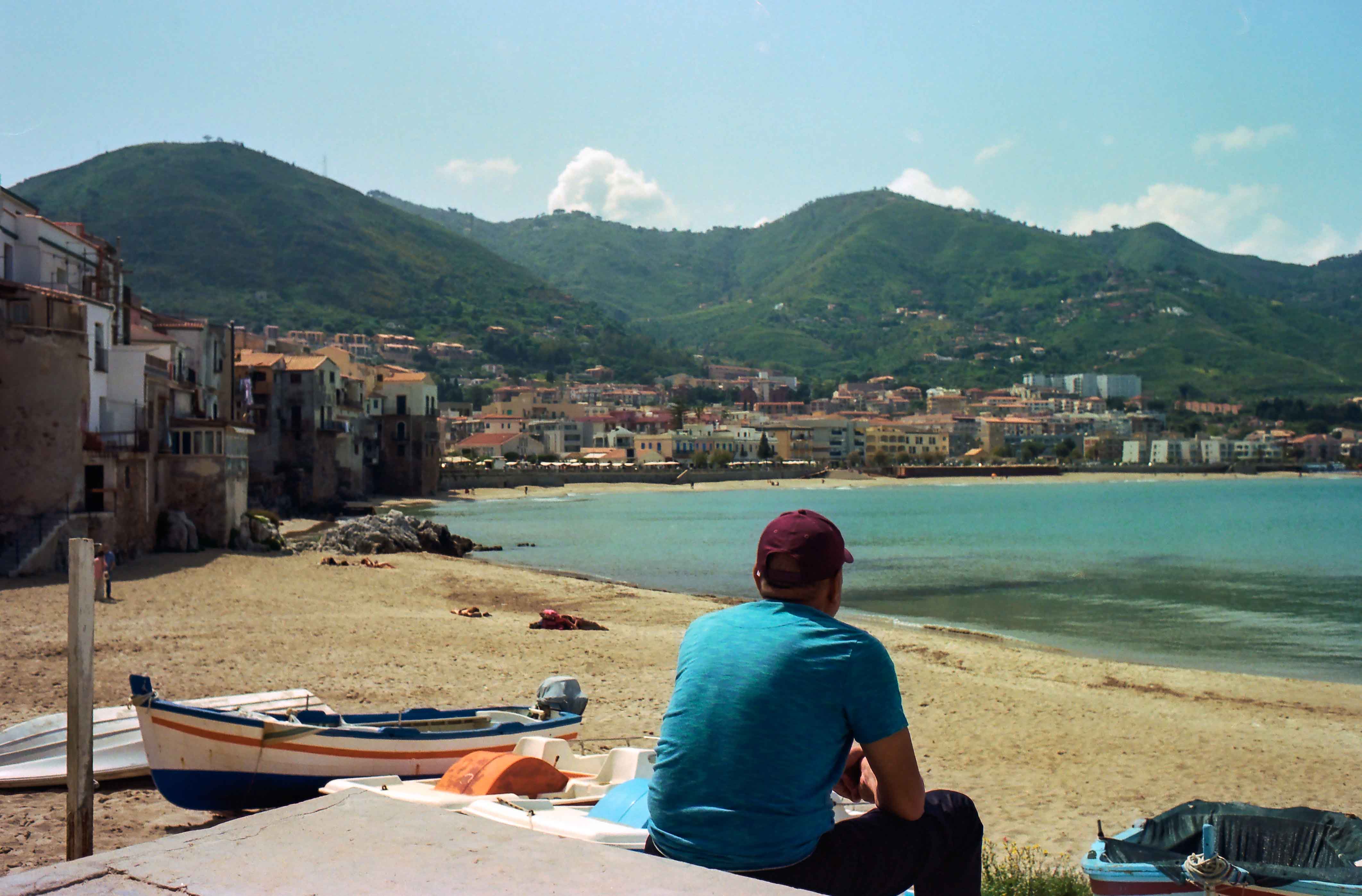 Persona con camiseta azul sentada frente a una playa con barcas y un pueblo entre montañas.
