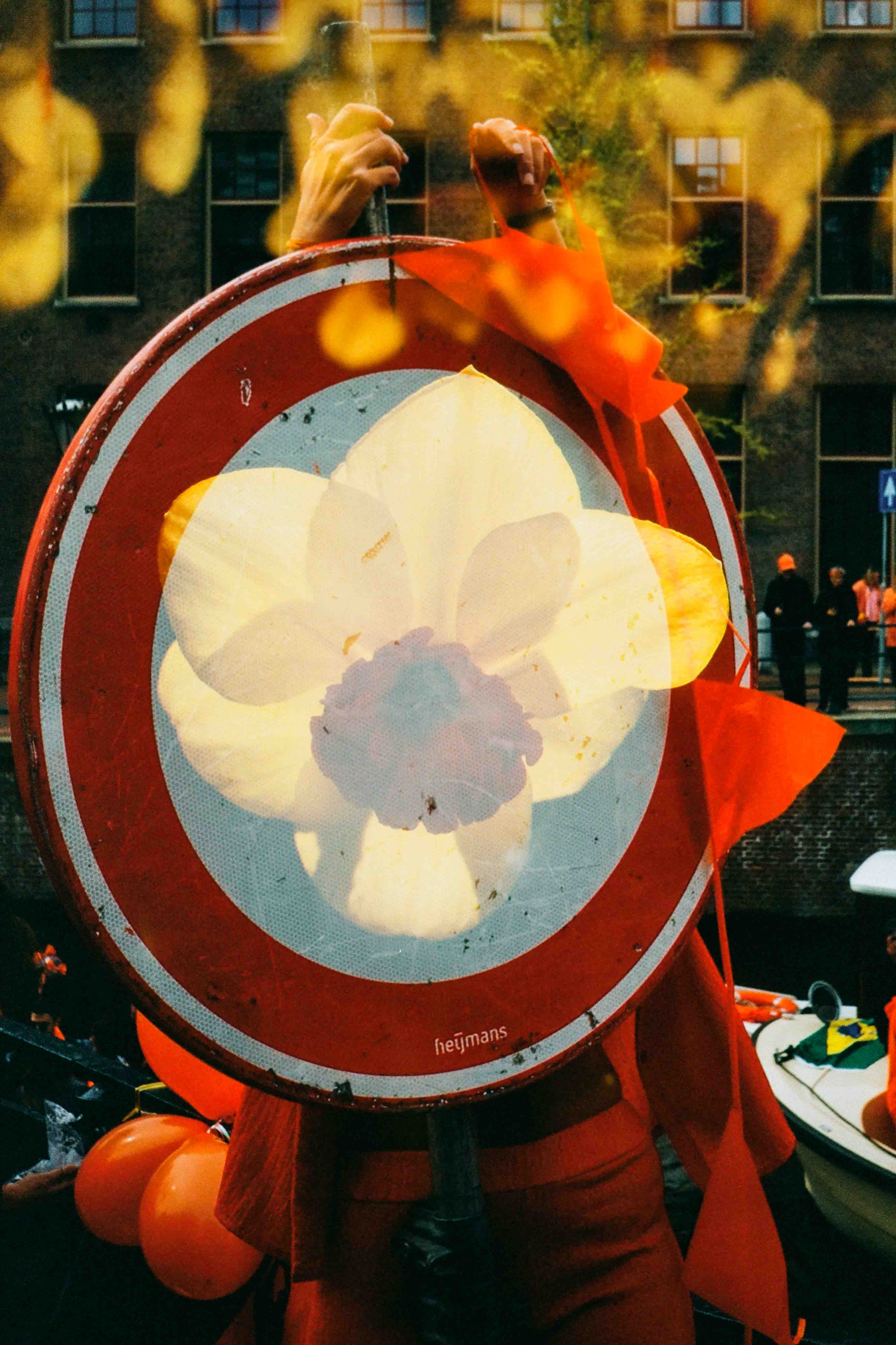 Persona de naranja detrás de un disco rojo y blanco, con una flor amarilla superpuesta y una cámara levantada sobre la cabeza.
