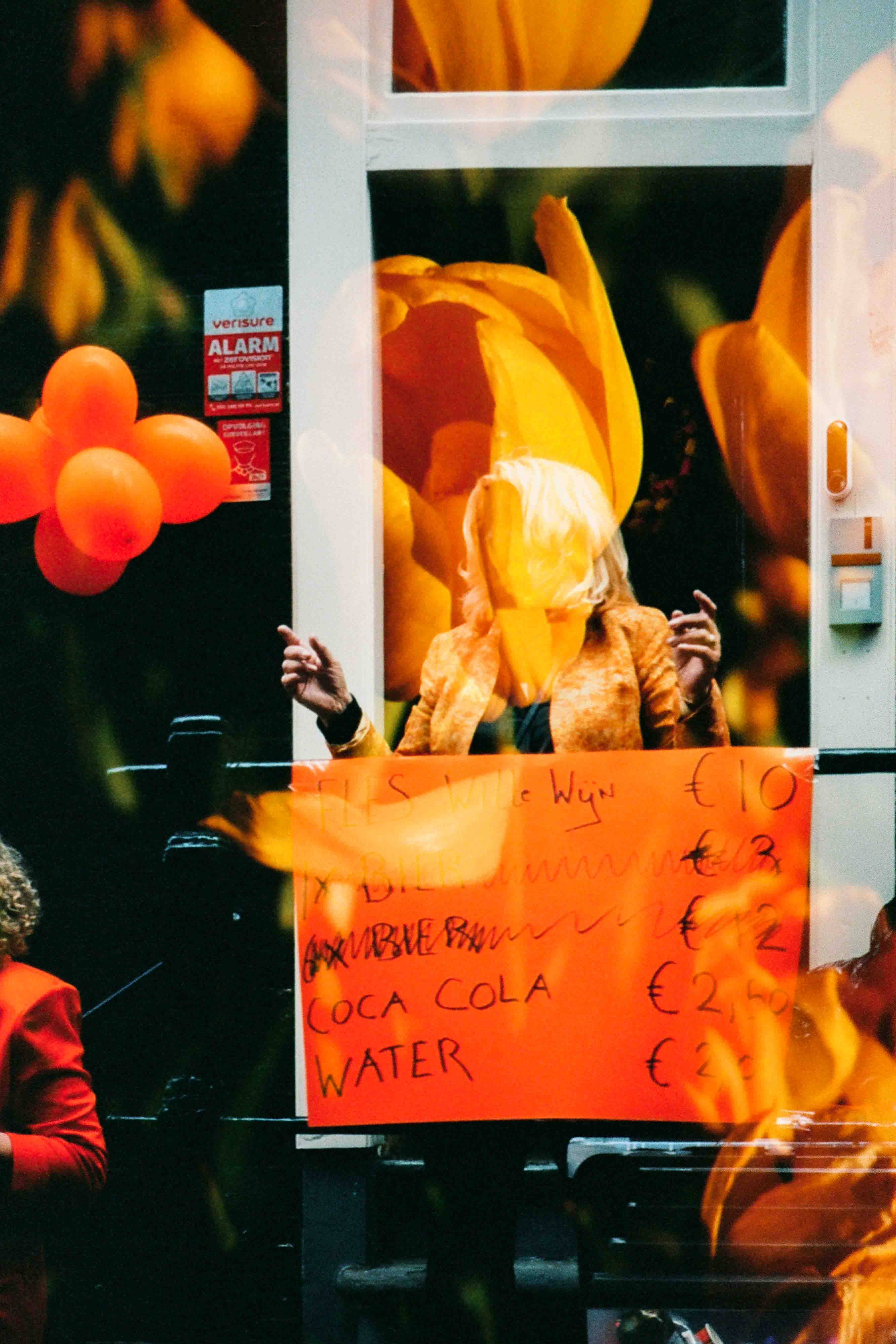 Ventana con globos naranjas y un cartel de bebidas que dice «COCA COLA» y «WATER», con flores superpuestas sobre una figura detrás.