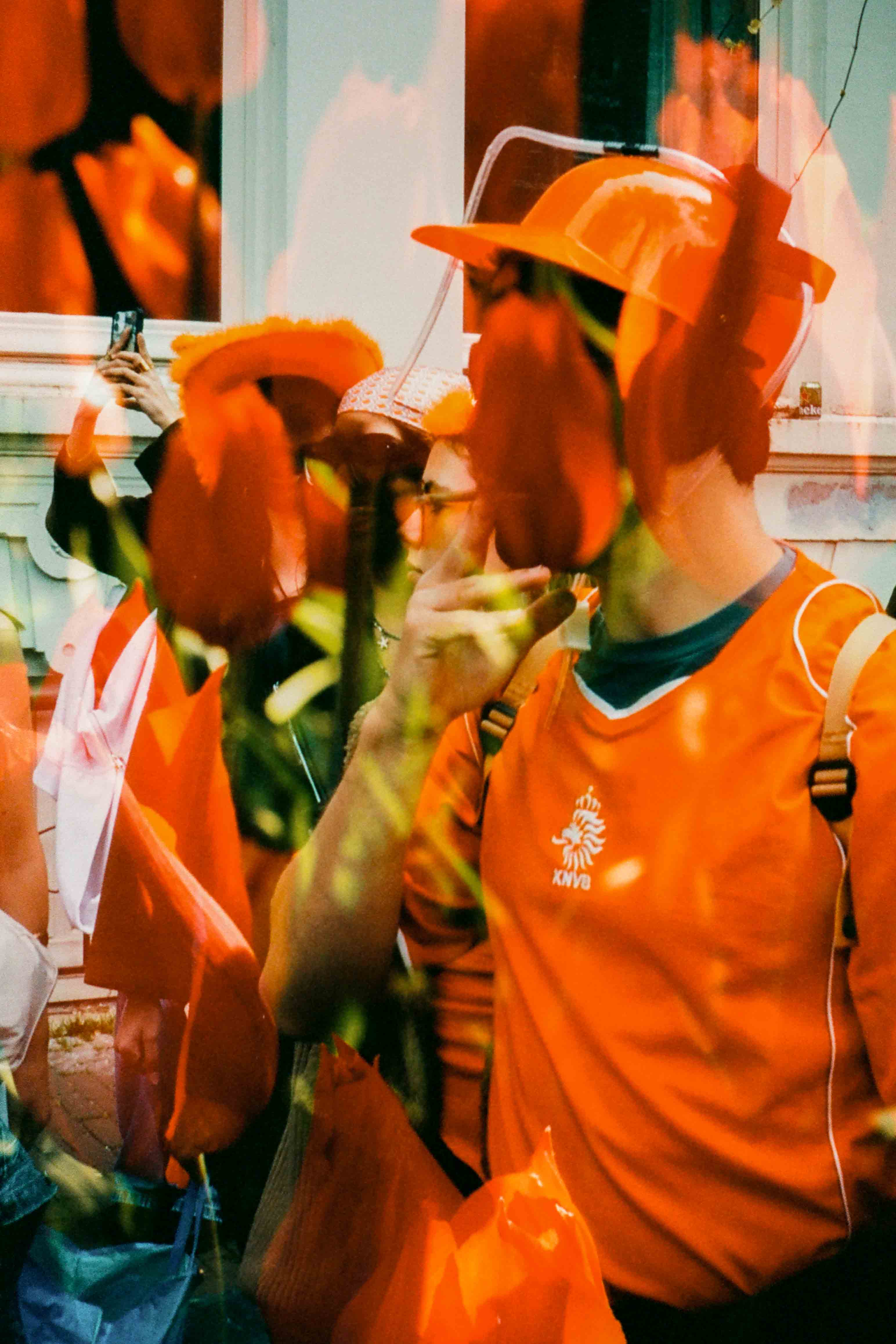 Persona con sombrero y camiseta naranjas, una mano cerca de la boca y tulipanes superpuestos sobre el rostro.