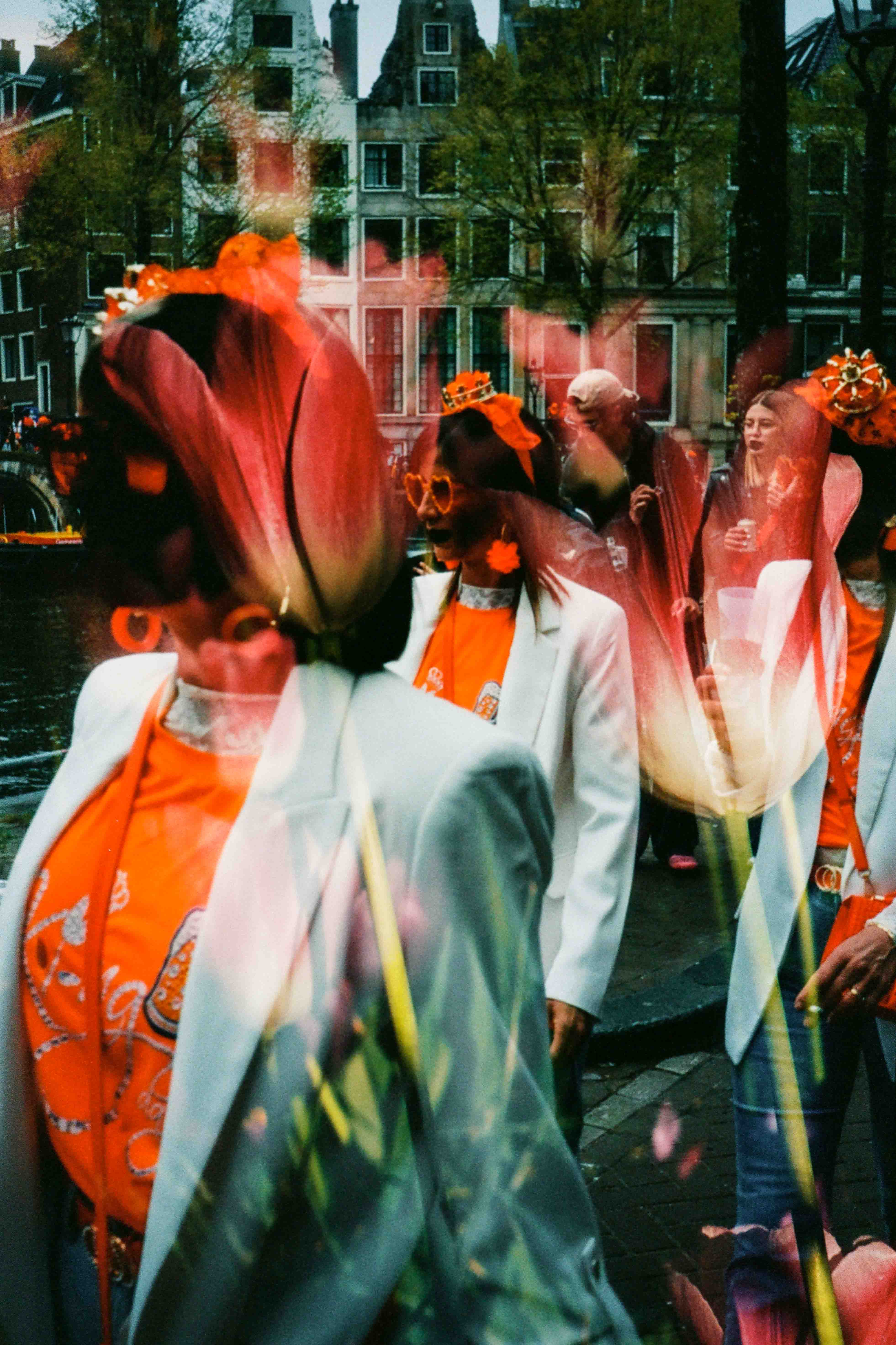 Personas con chaquetas blancas y camisetas naranjas en una calle, con tulipanes rojos y blancos sobre los rostros.